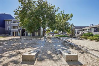 A concrete pathway leads through a sandy area with concrete blocks on either side, surrounded by trees and buildings.