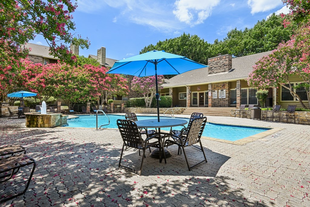 A poolside table with chairs and umbrellas is surrounded by a pool and trees. at The Glen, Lewisville, 75067