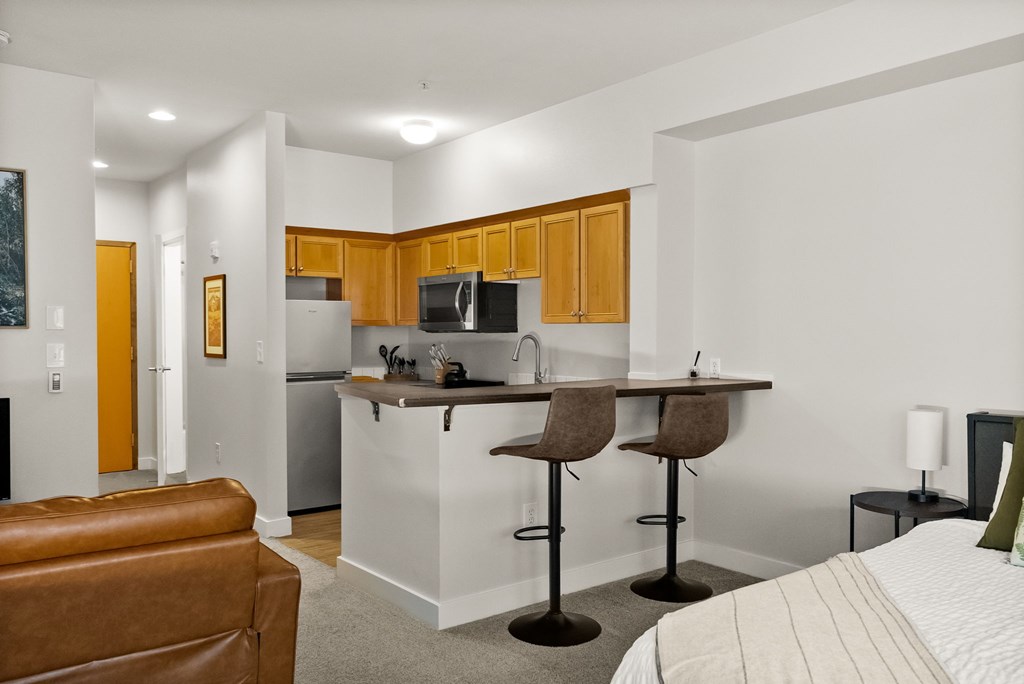 A living room with a brown leather couch and a kitchen area with a refrigerator, microwave, and cabinets.