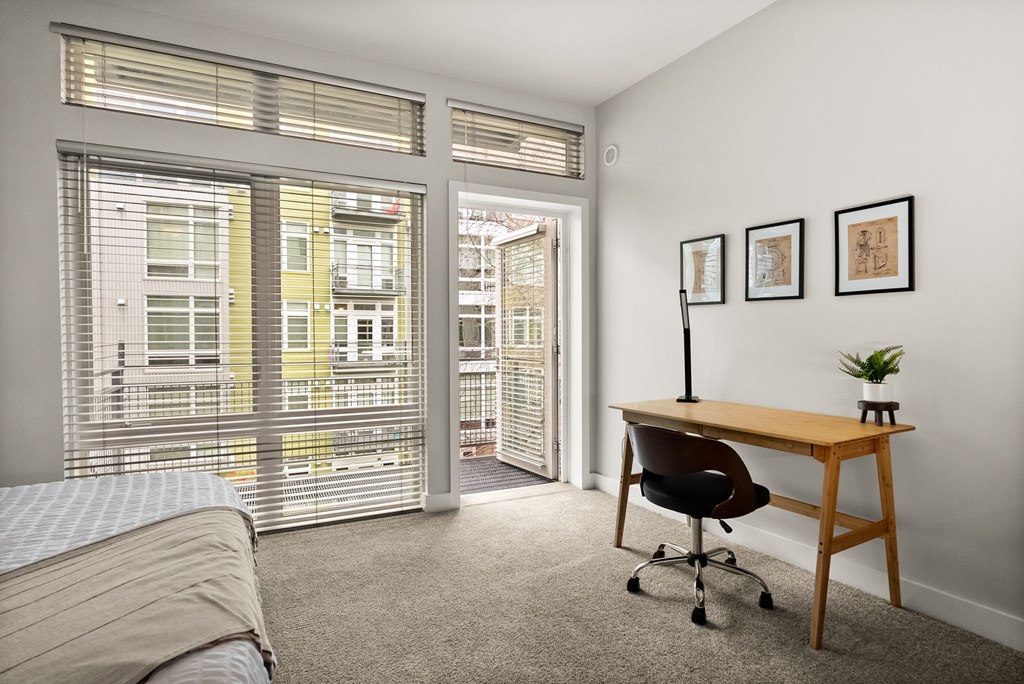 A room with a bed, desk, and chair with a window overlooking a balcony.