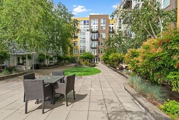 A patio with a table and chairs is surrounded by greenery and apartment buildings.