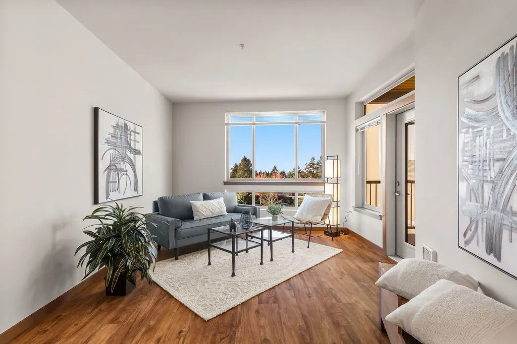 A living room with a grey couch, a white chair, a coffee table, and a large window with a view of trees.