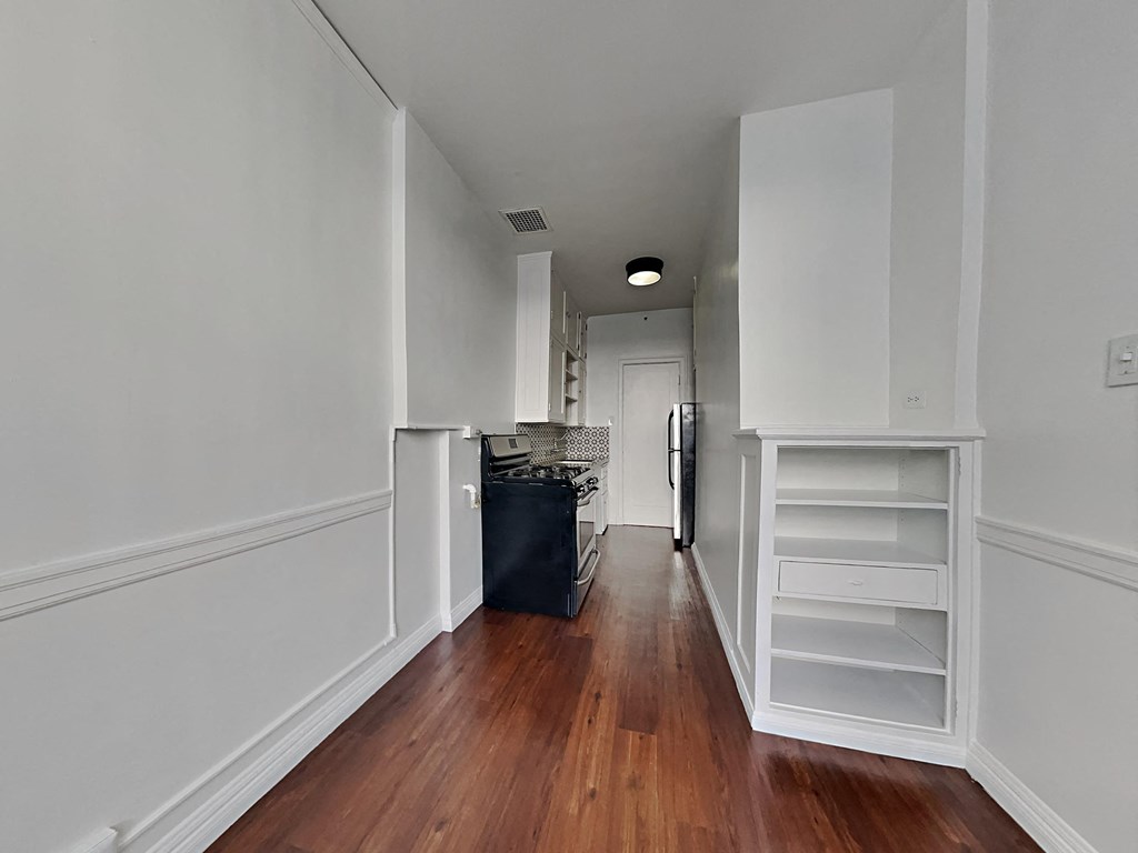 a living room with white walls and wood floors and a kitchen with white cabinets