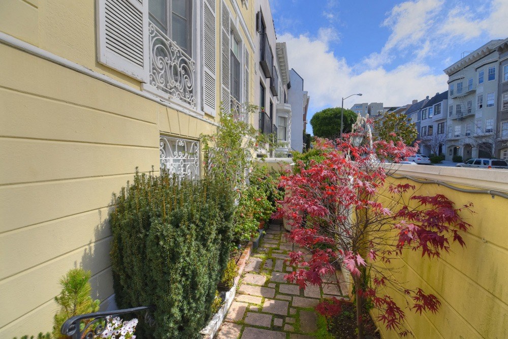 a side yard of a yellow house with plants and a sidewalk