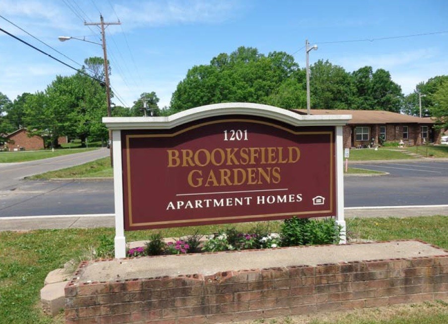 A sign for Brookfield Gardens Apartment Homes is displayed in front of a brick wall.