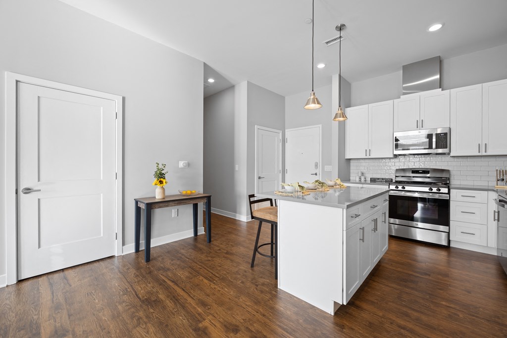 A modern kitchen with a breakfast bar, pendant lights, quartz countertops, stainless steel appliances, subway tile, and white cabinets near the entryway.