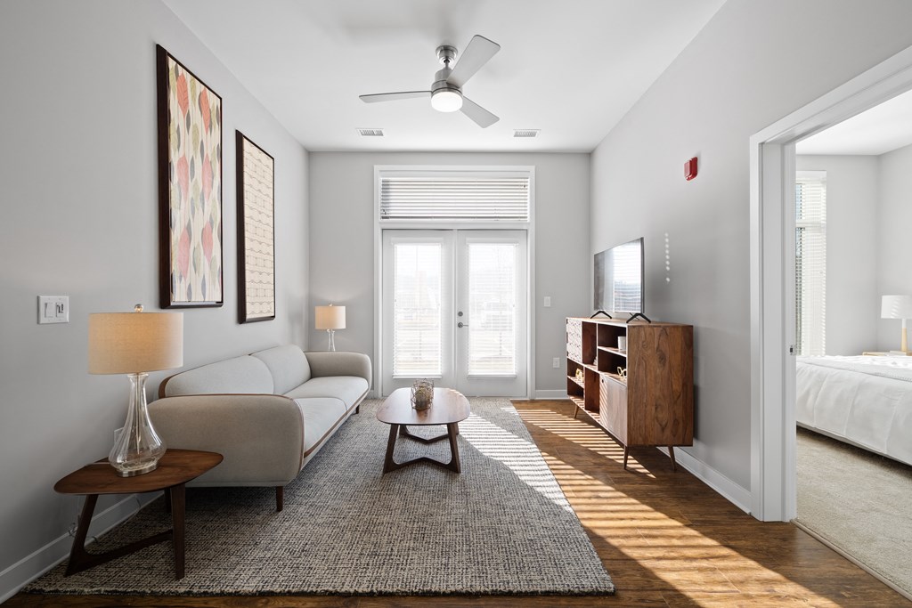 A living room with double doors to a patio providing natural light, hardwood-style flooring, a ceiling fan, and access to a bedroom.