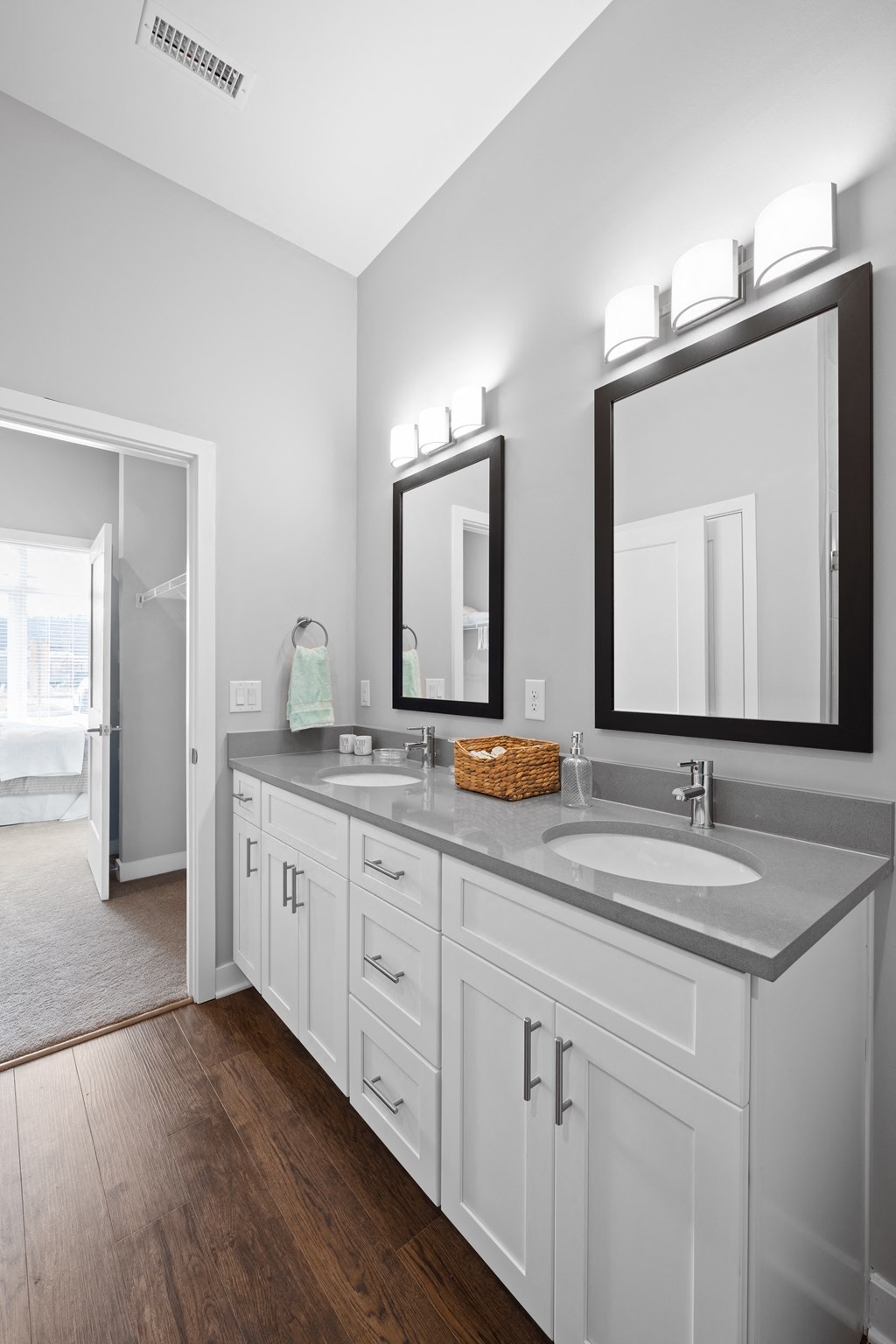 A bathroom with a double vanity, two mirrors, ample cabinet space, and hardwood-style flooring near a doorway to a walk-in closet and bedroom.