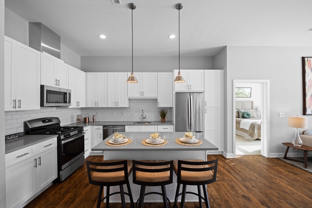 An L-shaped kitchen with a breakfast bar, two pendant lights, stainless steel appliances, hardwood-style flooring, and white cabinets.