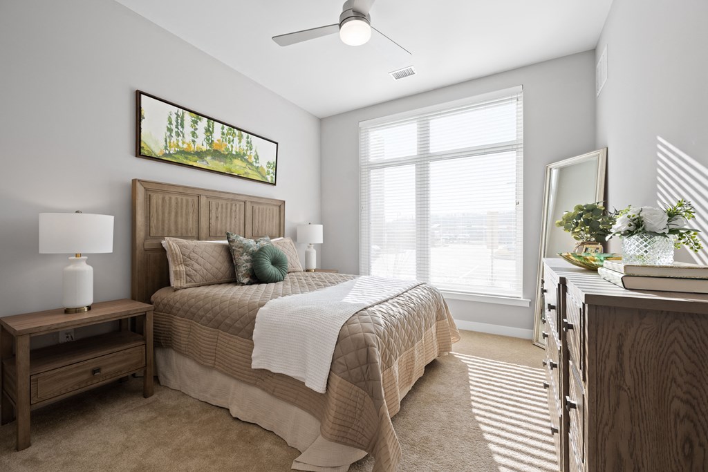A carpeted bedroom with ample natural light from a large window and a ceiling fan.