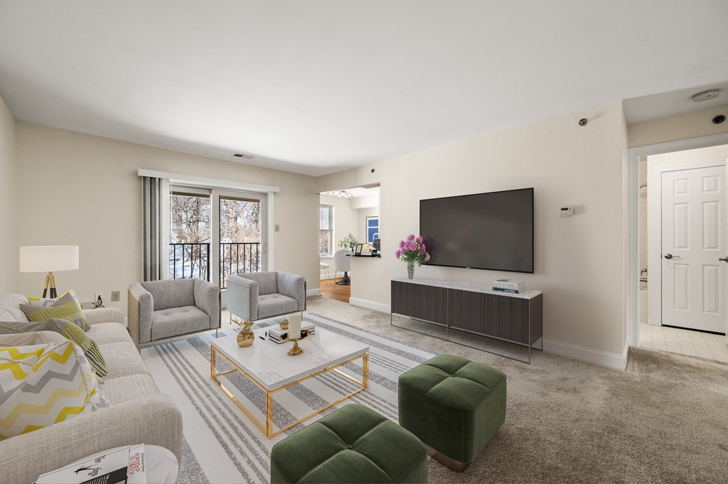 A living room with a grey sofa, a green ottoman, a coffee table, a television, and a window with a view of trees.