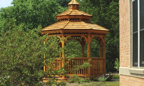 A wooden gazebo with a brown roof is surrounded by greenery.