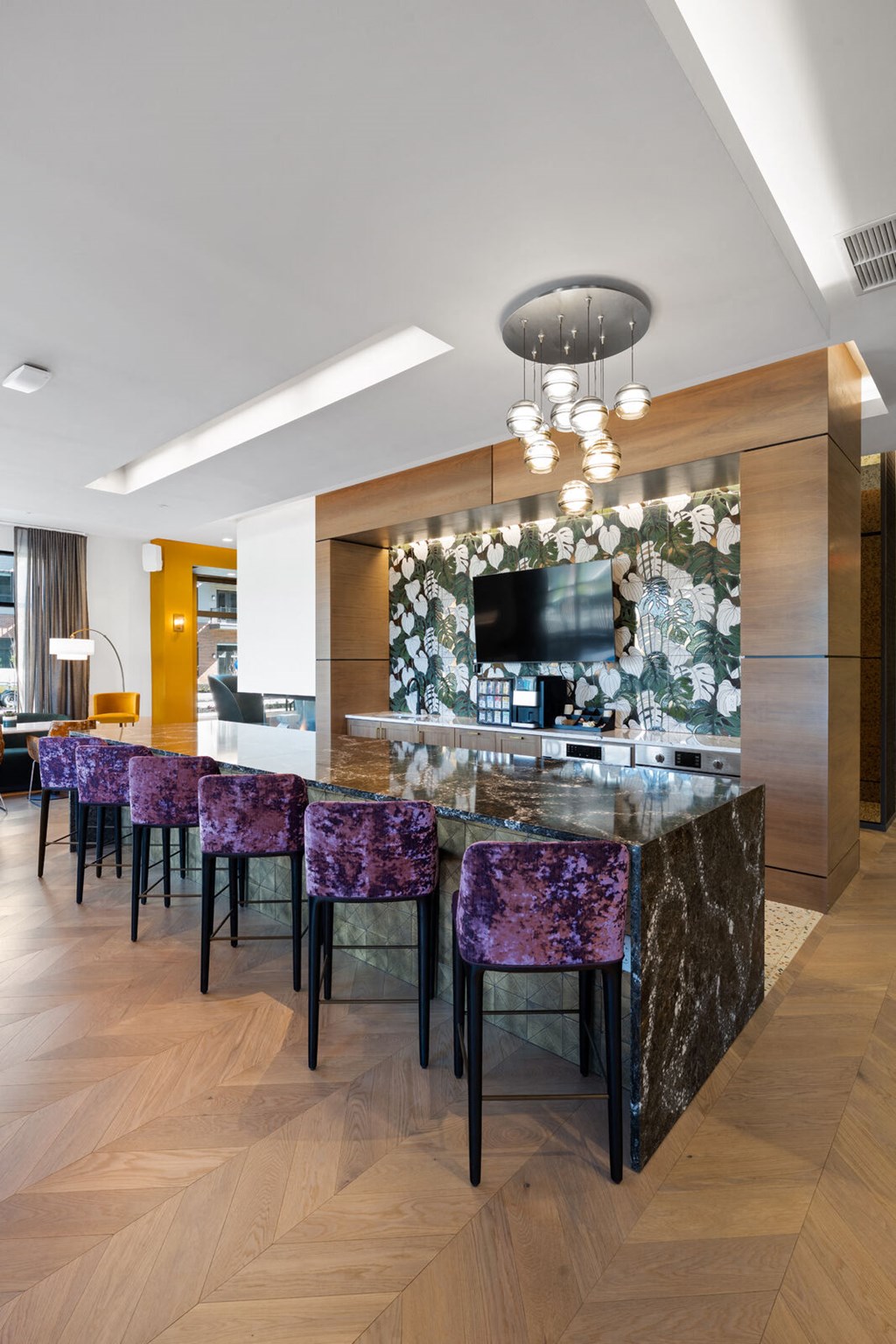 A community kitchen with a long bar, modern purple barstools, pendant chandelier, and a TV in a resident lounge.