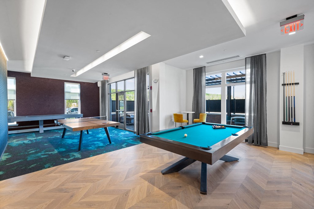 A clubhouse with a billiards table, a ping pong table, and a shuffleboard near doors leading to an outdoor community lounge.