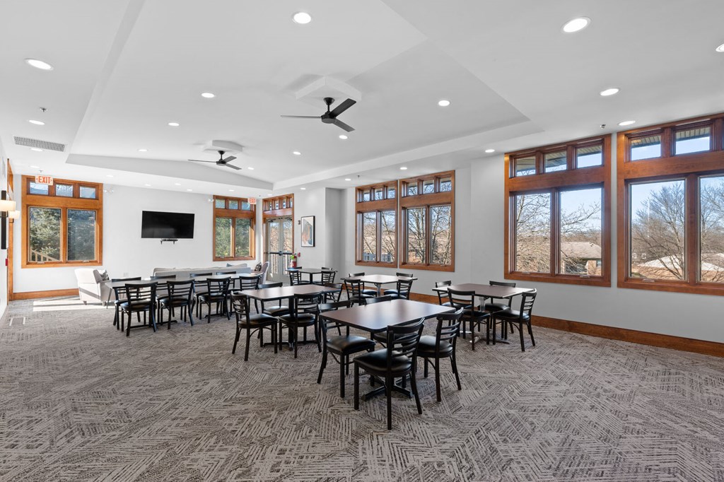 A clubhouse at New Kent Apartments in West Chester, PA, featuring tables, chairs, a TV, carpet flooring, recessed lighting, two ceiling fans, and large windows offering beautiful views
