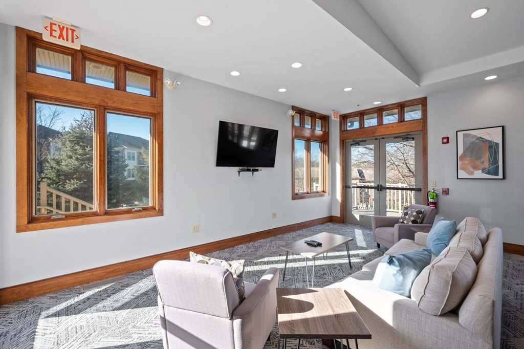 A community lounge at New Kent Apartments in West Chester, PA, with cozy seating, coffee tables, a TV, and windows offering views of the outdoor green space
