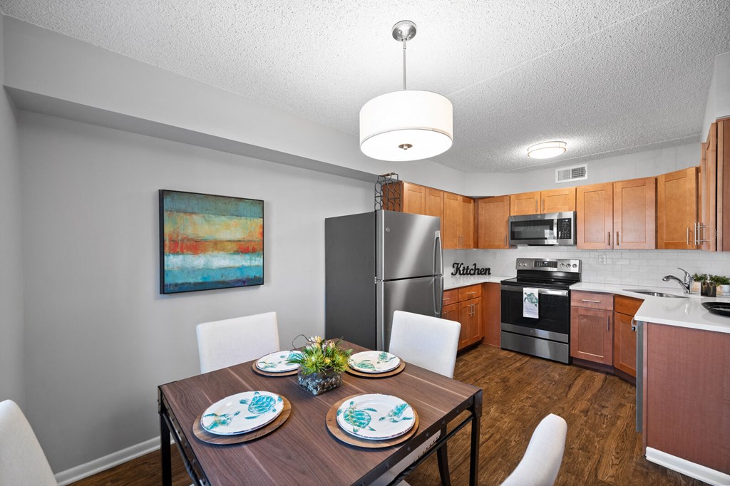 A dining room with a pendant light near a U-shaped kitchen with stainless steel appliances, hardwood style flooring, and a subway tile backsplash.