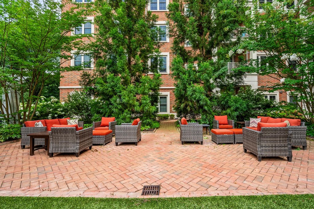 A patio with red cushions and grey wicker furniture is surrounded by trees and a brick building.