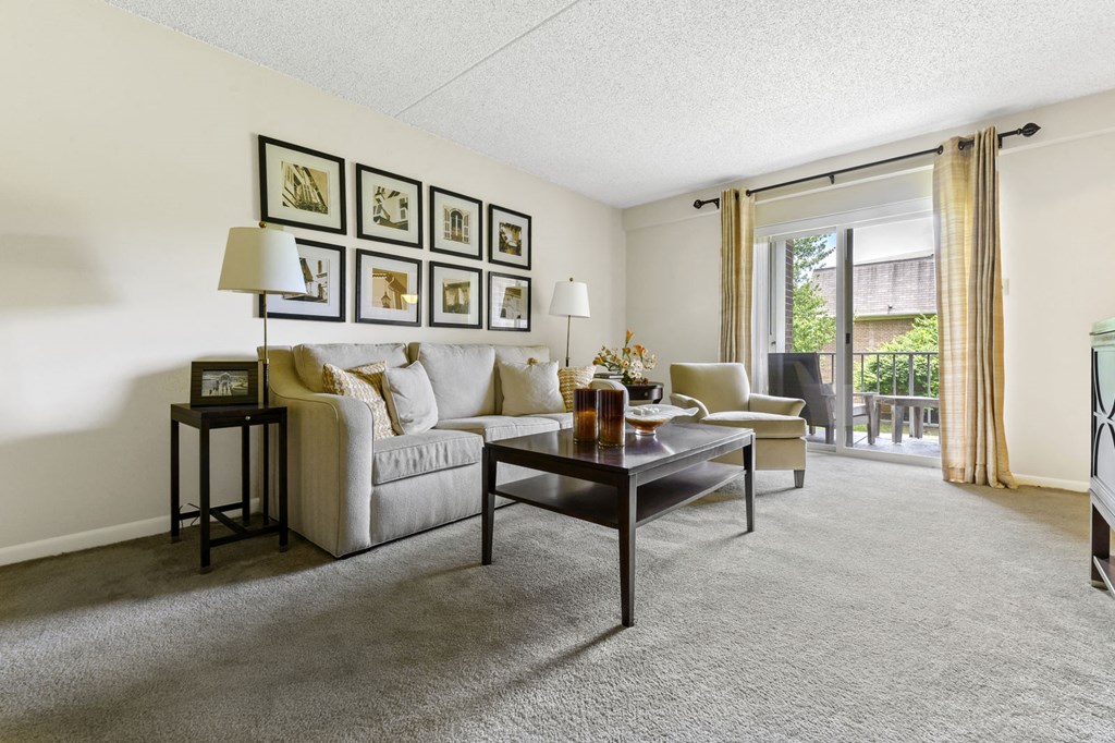 A spacious living room at New Kent Apartments in West Chester, PA, featuring carpet flooring, sliding doors leading to a private balcony, and plenty of natural light