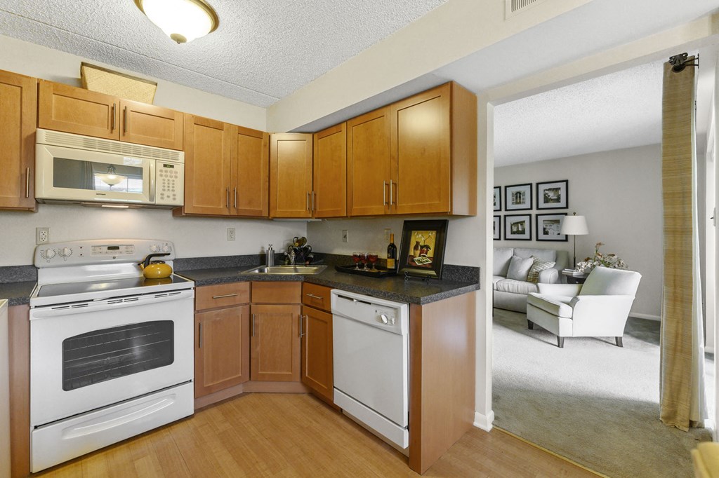 An L-shaped kitchen with white appliances, hardwood-style flooring, and open entryway to a carpeted living room.