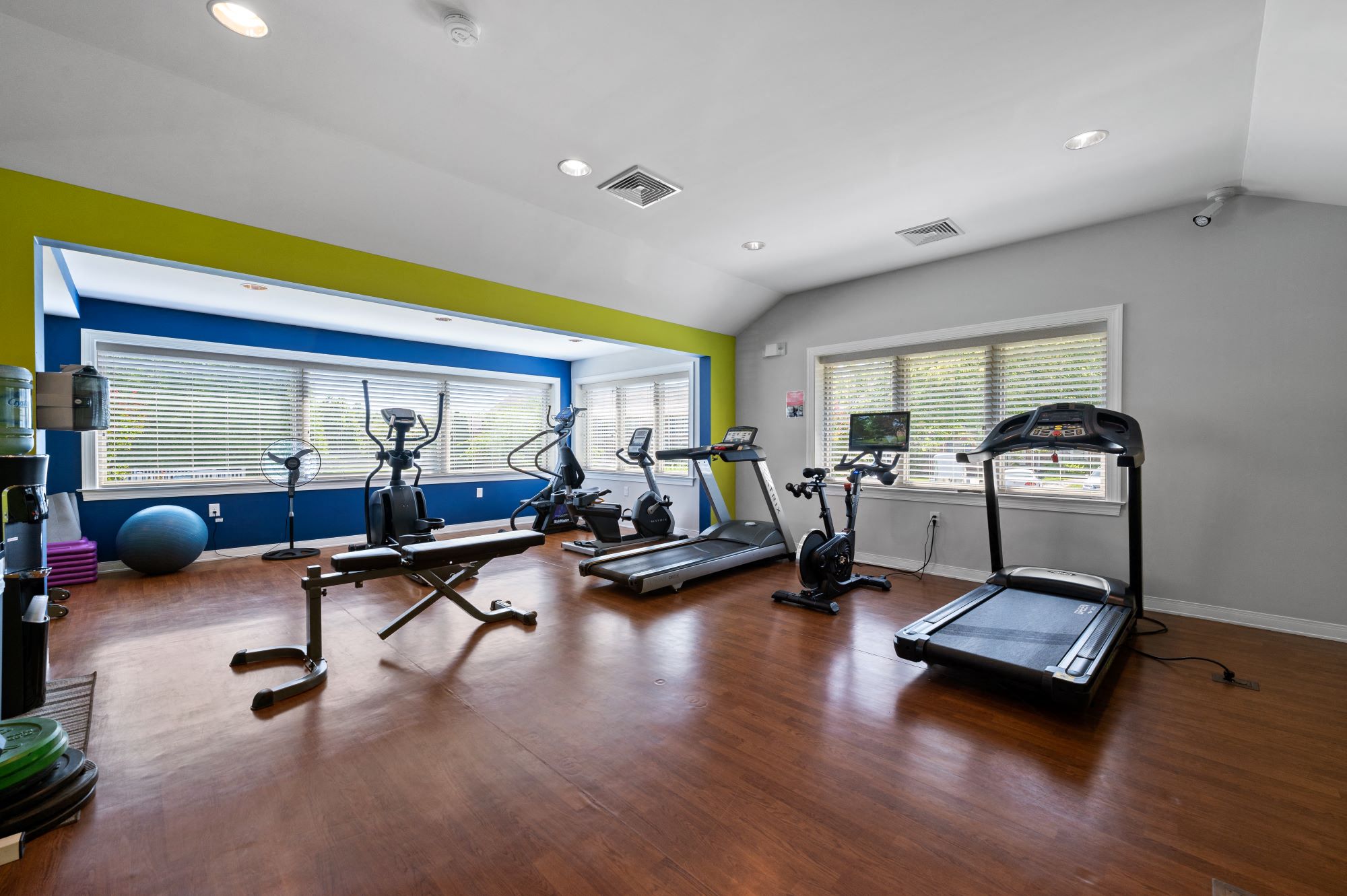 A spacious fitness center with beautiful hardwood flooring, large windows, cardio and weight equipment, a filtered water station, and large windows.