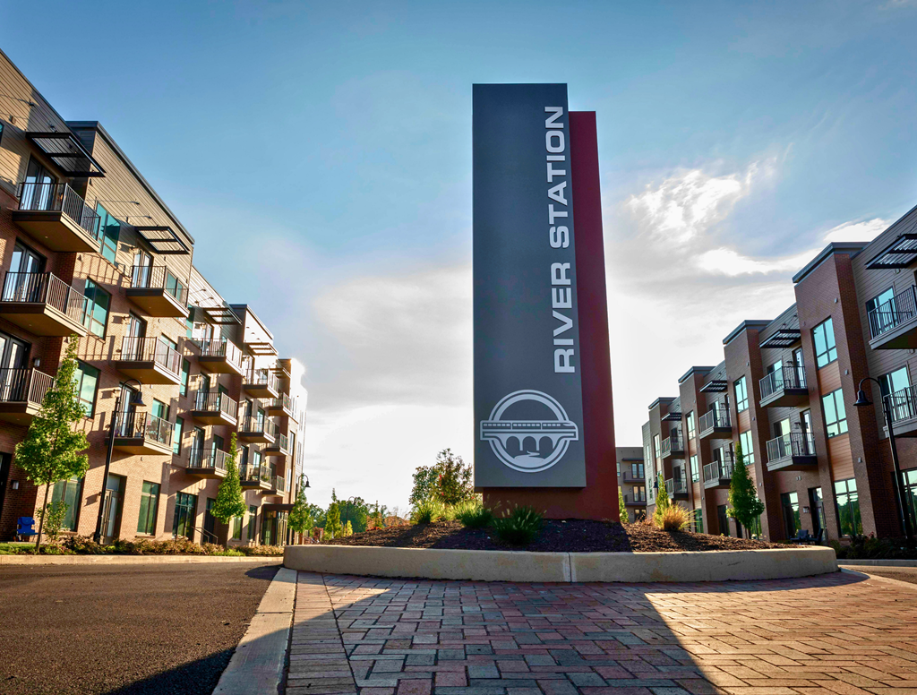 A River Station sign in front of modern apartment buildings with balconies and trees lining a street.