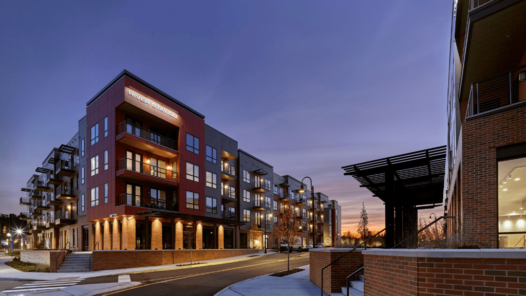 A corner street view of River Station's apartment buildings at sunset with trees and the capitol building in the background.