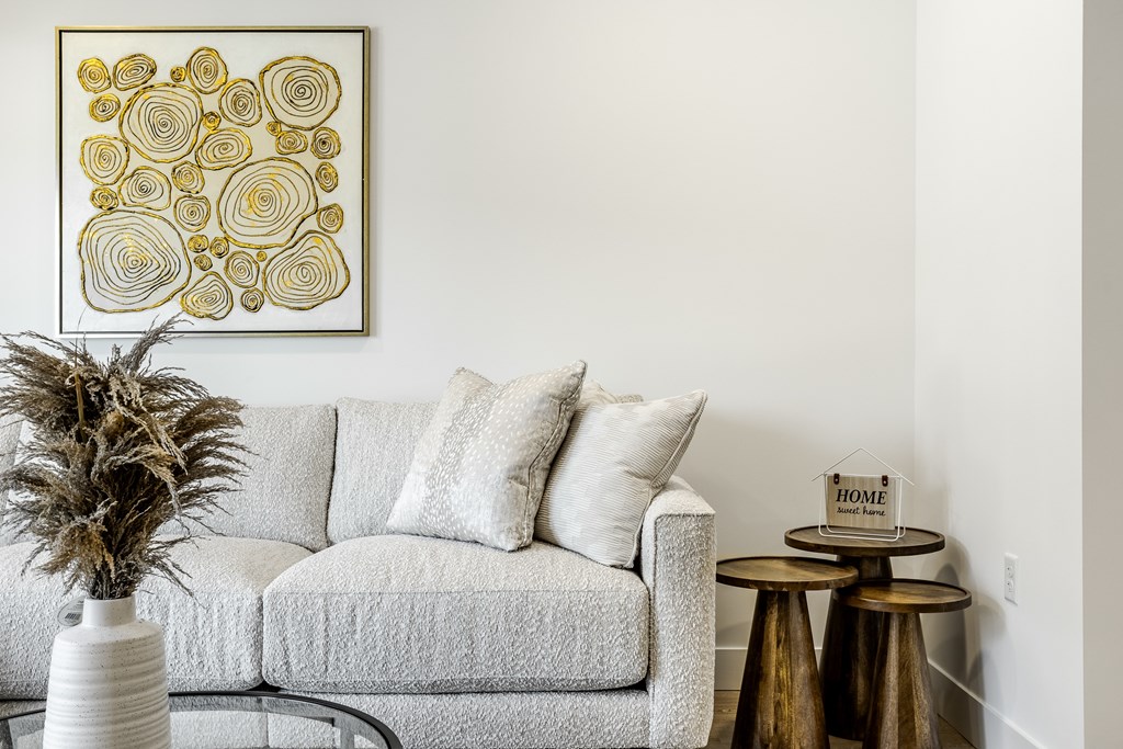 A living room with a grey couch, a small table with a house sign, and a framed artwork on the wall.