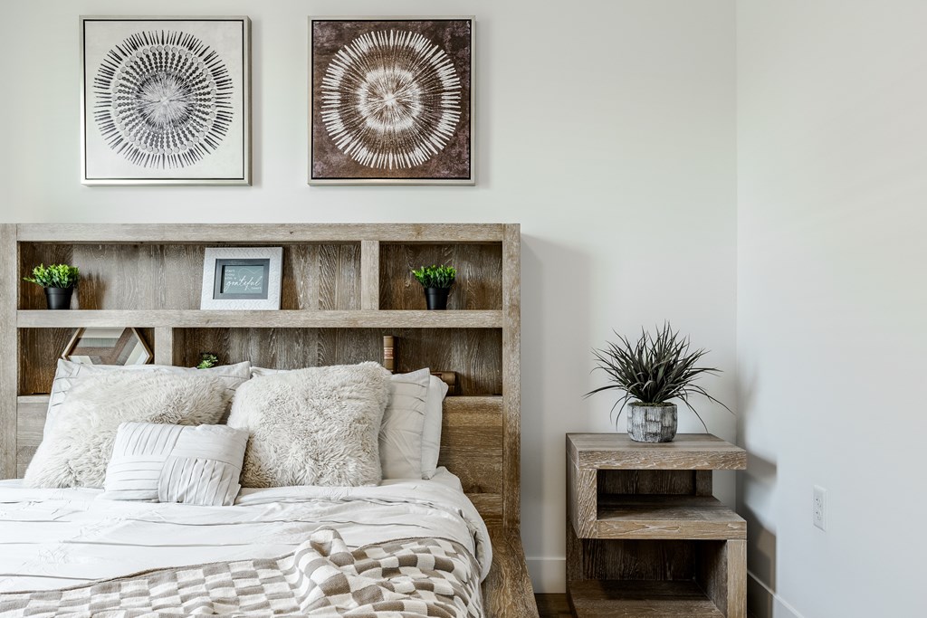 A bedroom with a bed, a wooden shelf with a plant, and two wall art pieces at Prairie Encore, Dardenne Prairie, MO 63368