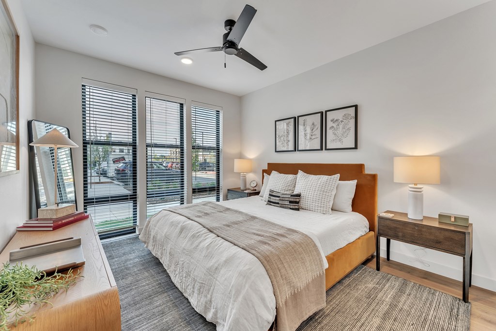 A bedroom with a large bed, a ceiling fan, and a view of the street through the windows.