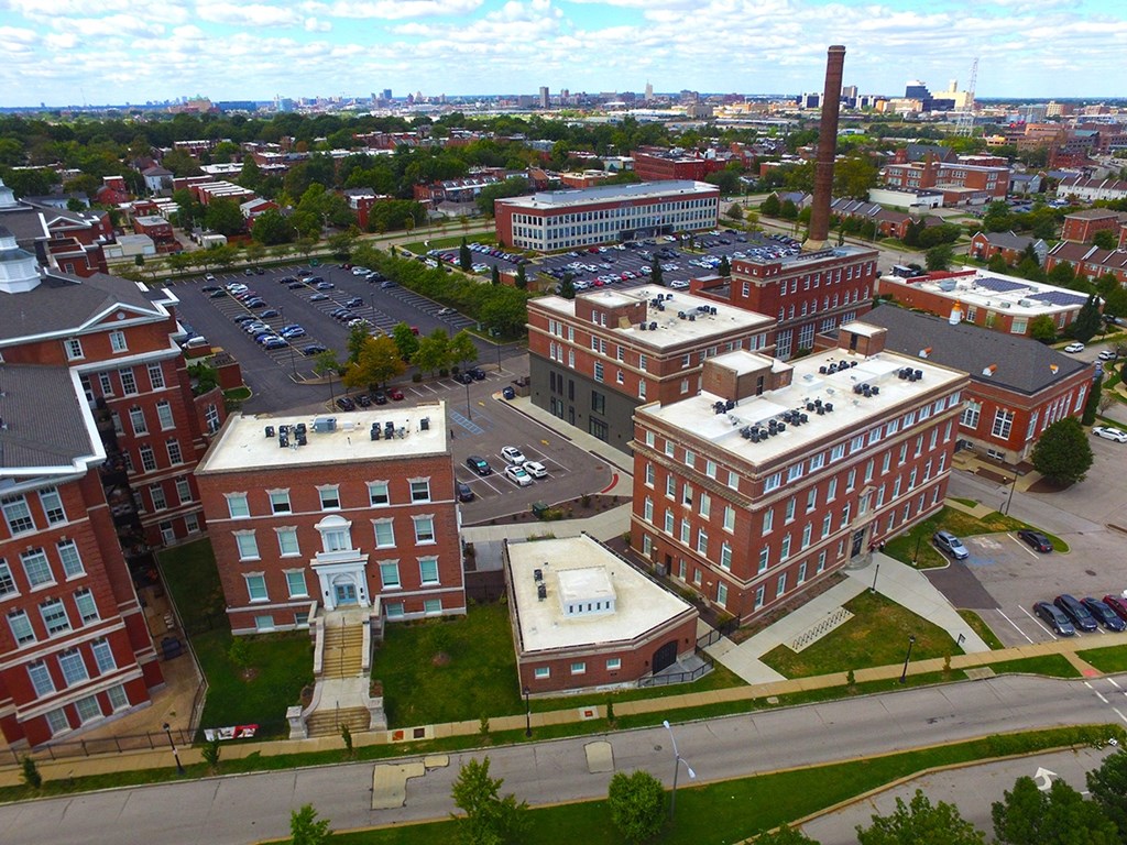 A large brick building with a tall chimney is surrounded by other buildings and a parking lot.