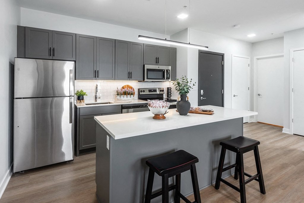 model kitchen with stainless steel appliances, plank flooring, kitchen island, white quartz countertops, gray cabinetry and white tile backsplash at Steelcote Square, St. Louis, 63103