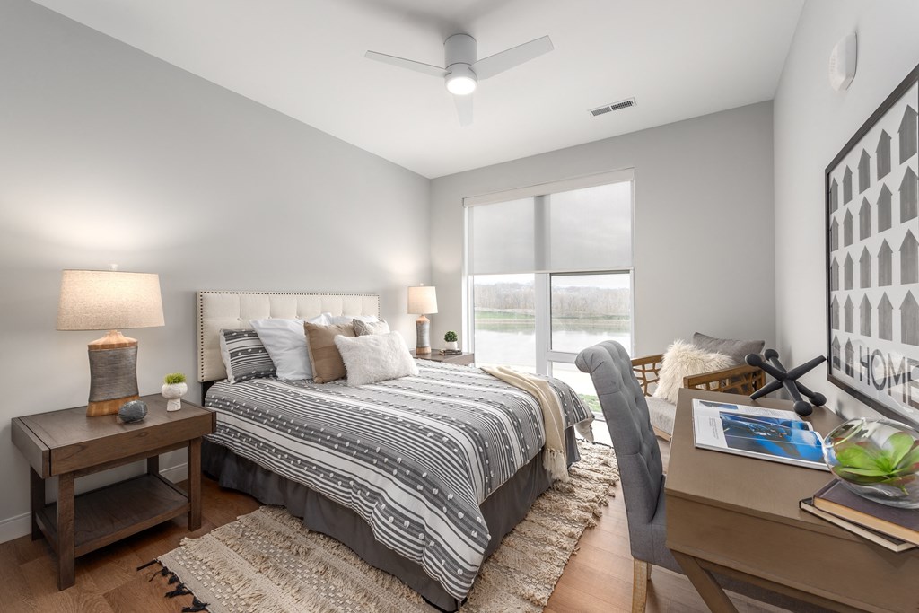 model bedroom with plank flooring, ceiling fan, large windows, large bedroom with nightstand, accent chair and desk at The Flats & Terraces at Wildhorse Village, Chesterfield Missouri