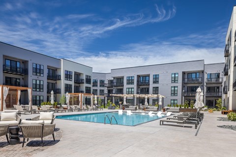A large swimming pool surrounded by lounge chairs and umbrellas in front of a modern apartment complex.