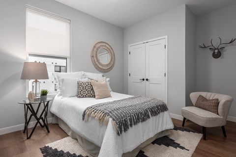 2nd bedroom with rich plank flooring, oversized window with custom shade, full size bed with nightstand and arm chair at The Flats & Terraces at Wildhorse Village, Chesterfield