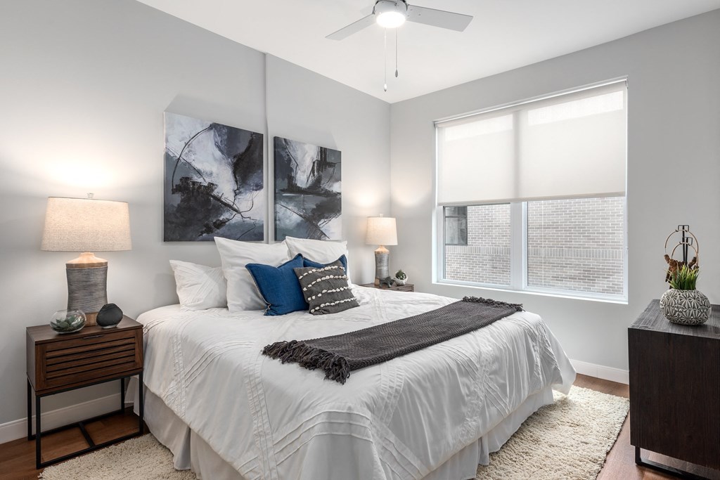 main bedroom with rich plank flooring, oversized windows with custom shades, ceiling fan, large bed, nightstands and accent table at The Flats & Terraces at Wildhorse Village, Chesterfield Missouri