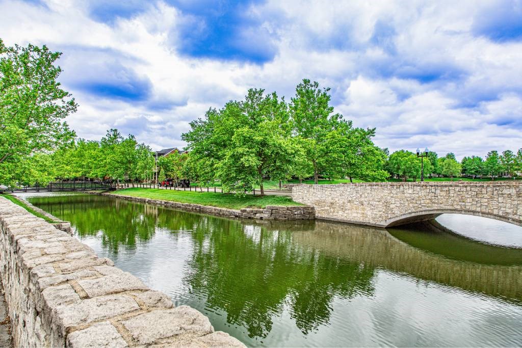 A stone bridge over a green water body with trees in the background.