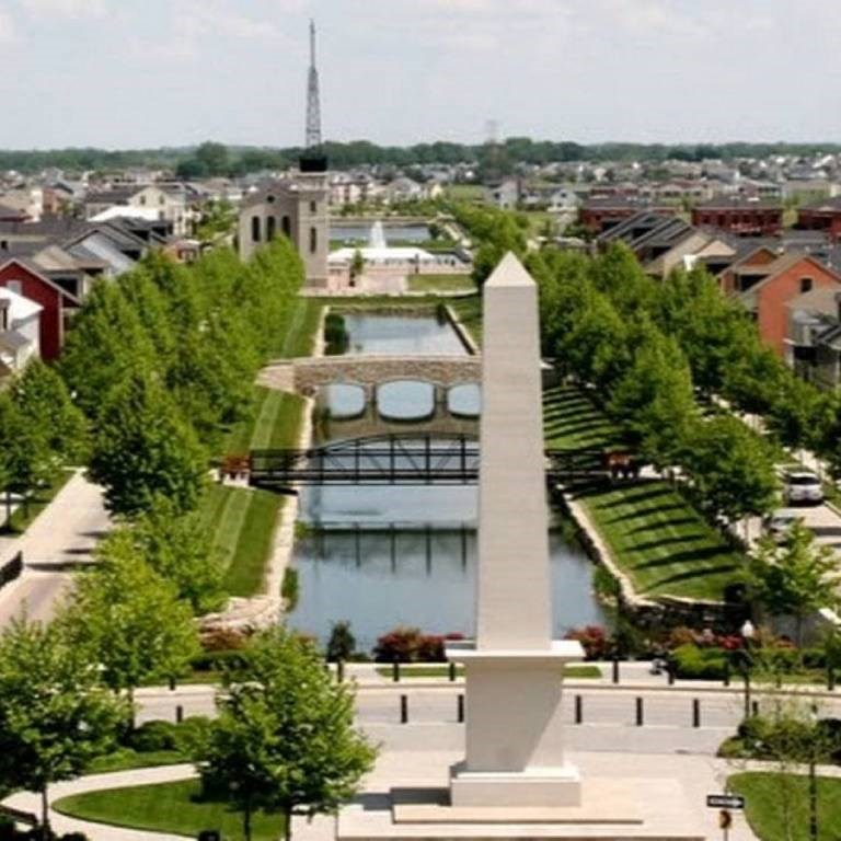 A monument stands in the middle of a park with a lake and a bridge in the background.