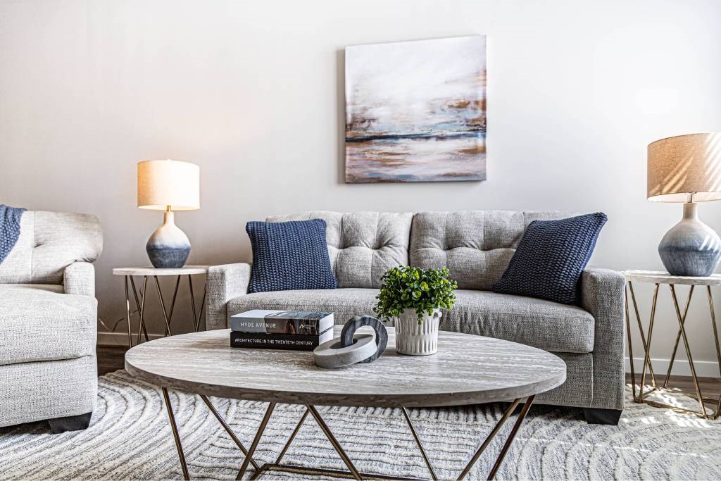 A living room with a grey couch, a grey armchair, a grey coffee table, a grey rug, a white side table, a white floor lamp, a white floor lamp, a white floor lamp, a white floor lamp, a
