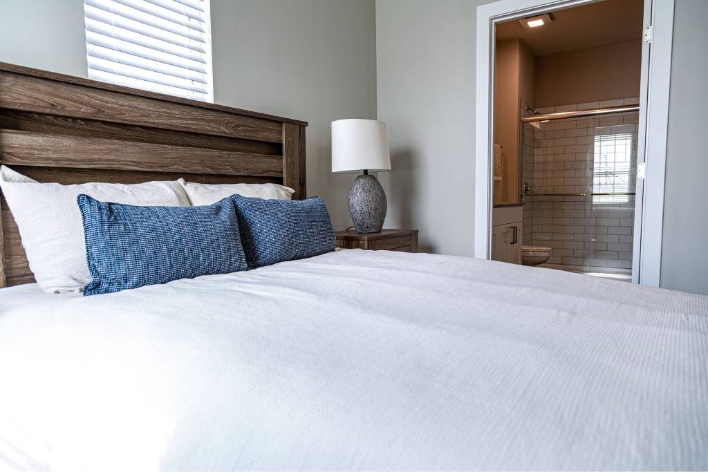 A bed with white sheets and blue pillows is in the foreground with a lamp on a side table.