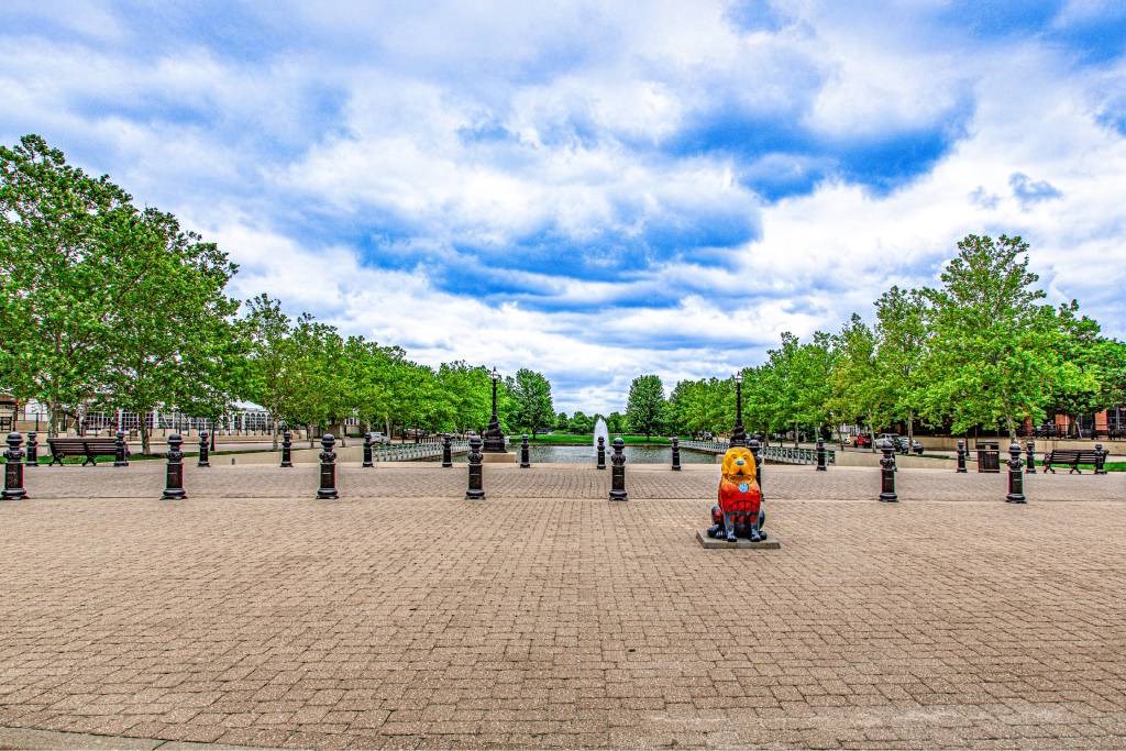 A person in a red and yellow outfit is standing in the middle of a brick-paved area with trees and bollards in the background.