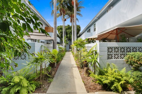 A concrete walkway leads between two white buildings with a fence and green plants on either side.