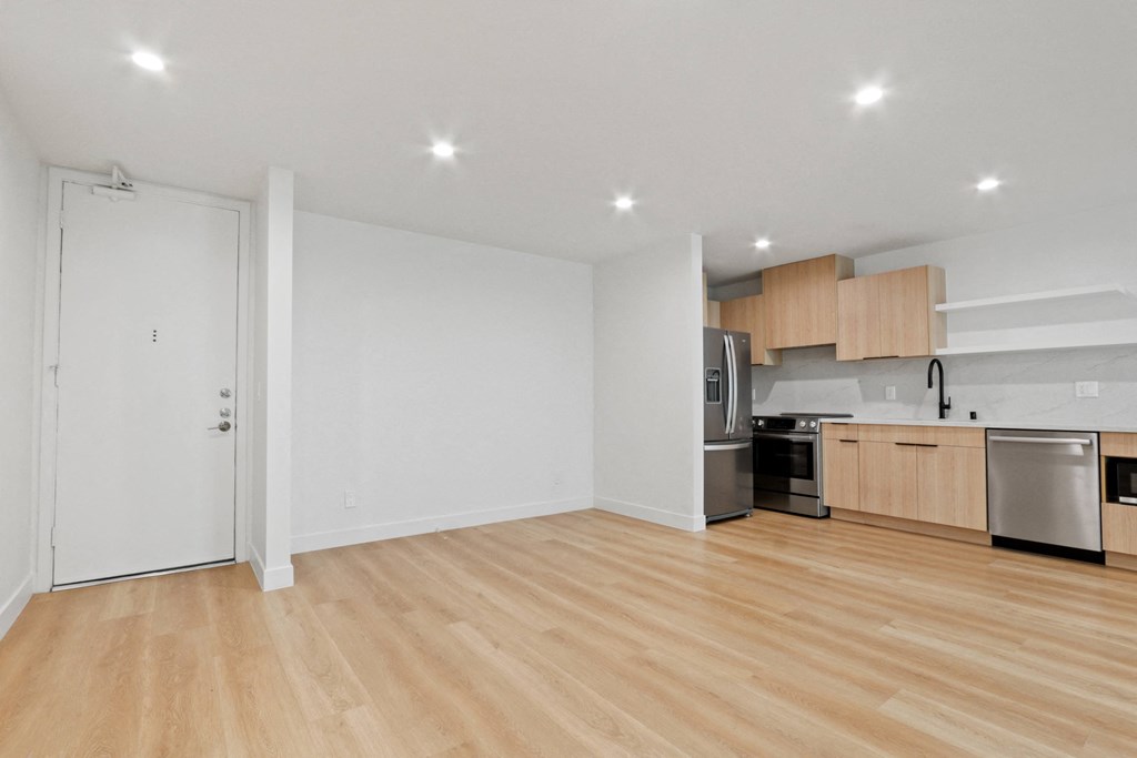 an empty living room with wood flooring and a kitchen with stainless steel appliances