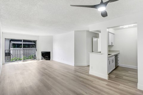 A spacious room with a ceiling fan and light, hardwood floors, and a kitchenette with white cabinets.