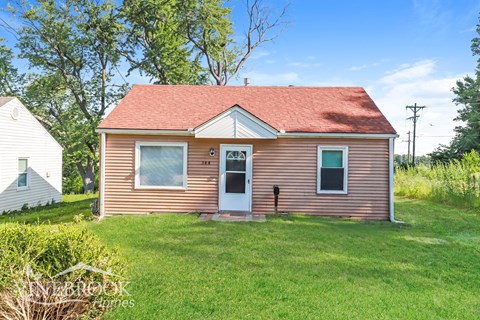 A small house with a red roof and a white door is surrounded by green grass.