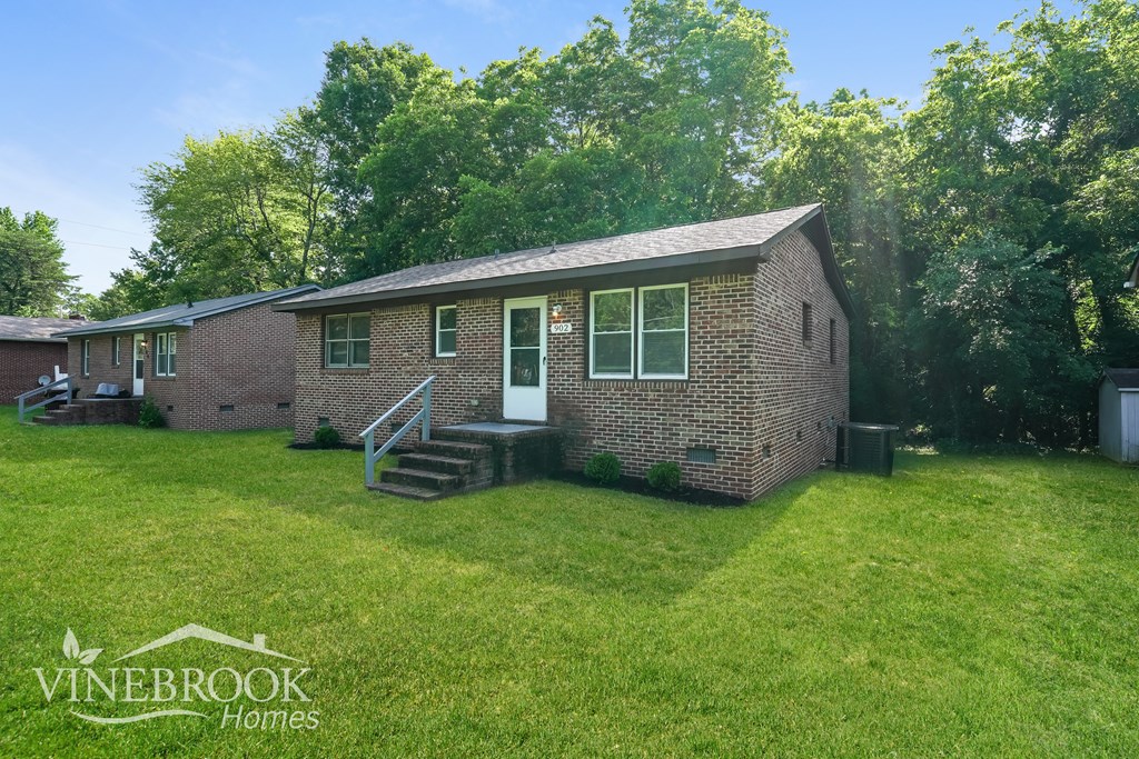 A brick house with a white door and a small porch is surrounded by green grass and trees.