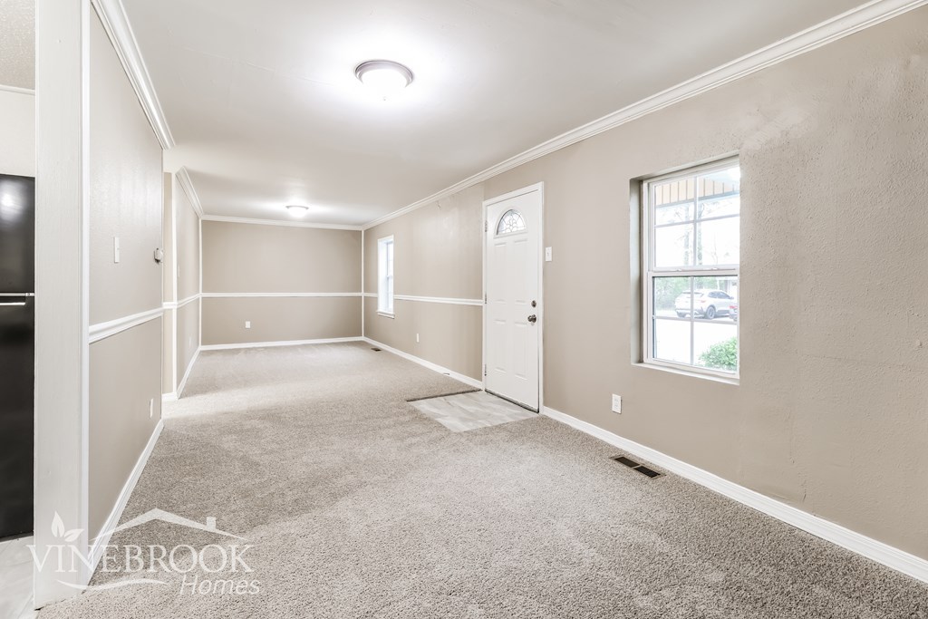 A spacious room with beige walls and carpet flooring, featuring a window with a view of greenery outside.