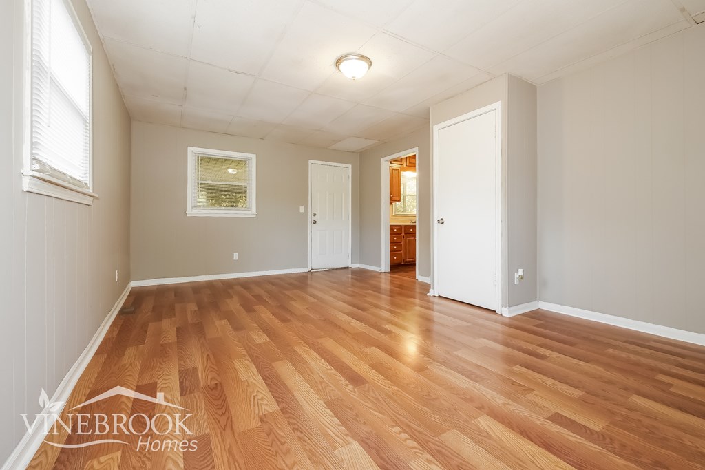 A room with wooden floors and white walls, with a window on the left and a door on the right.
