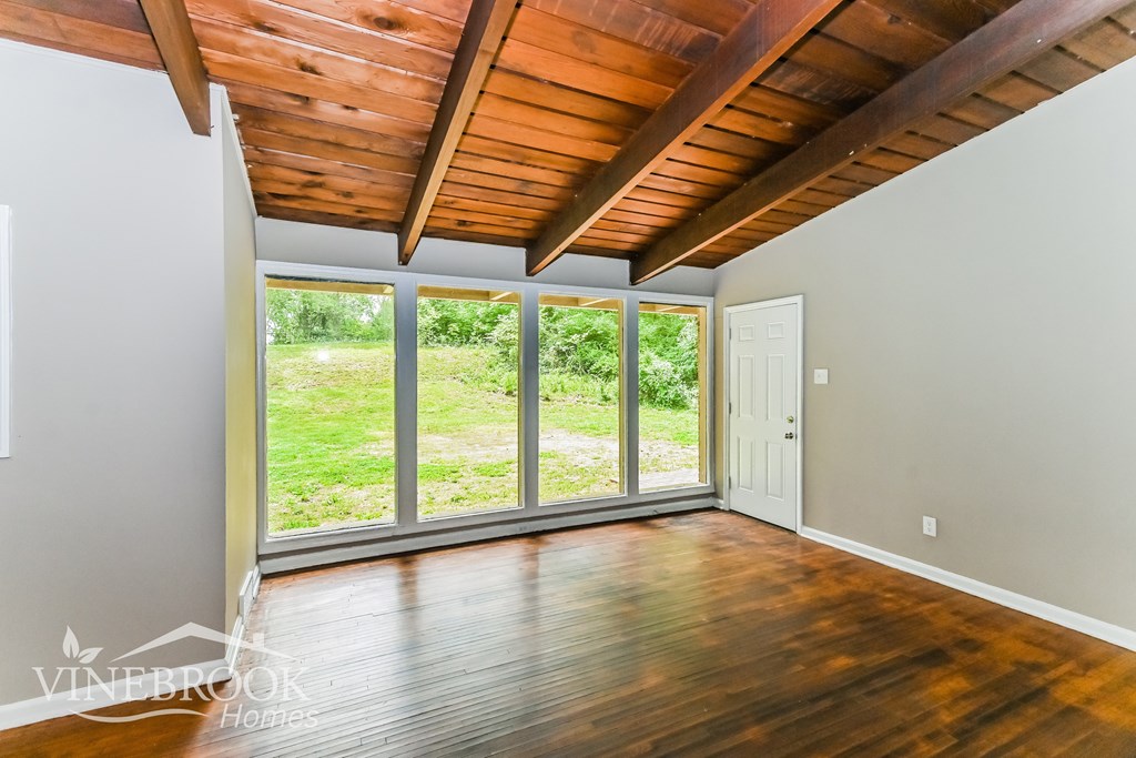 A room with a wooden ceiling and floor with a white wall and a door leading to a grassy area.