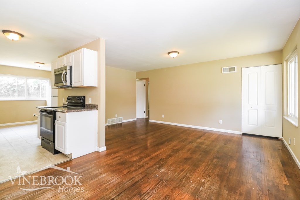 A spacious living room with wood flooring and a kitchen area in the background.